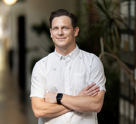 A nimble person in a white shirt with cross patterns stands with arms crossed, wearing a smartwatch, in a corridor adorned with plants and a blurred background.