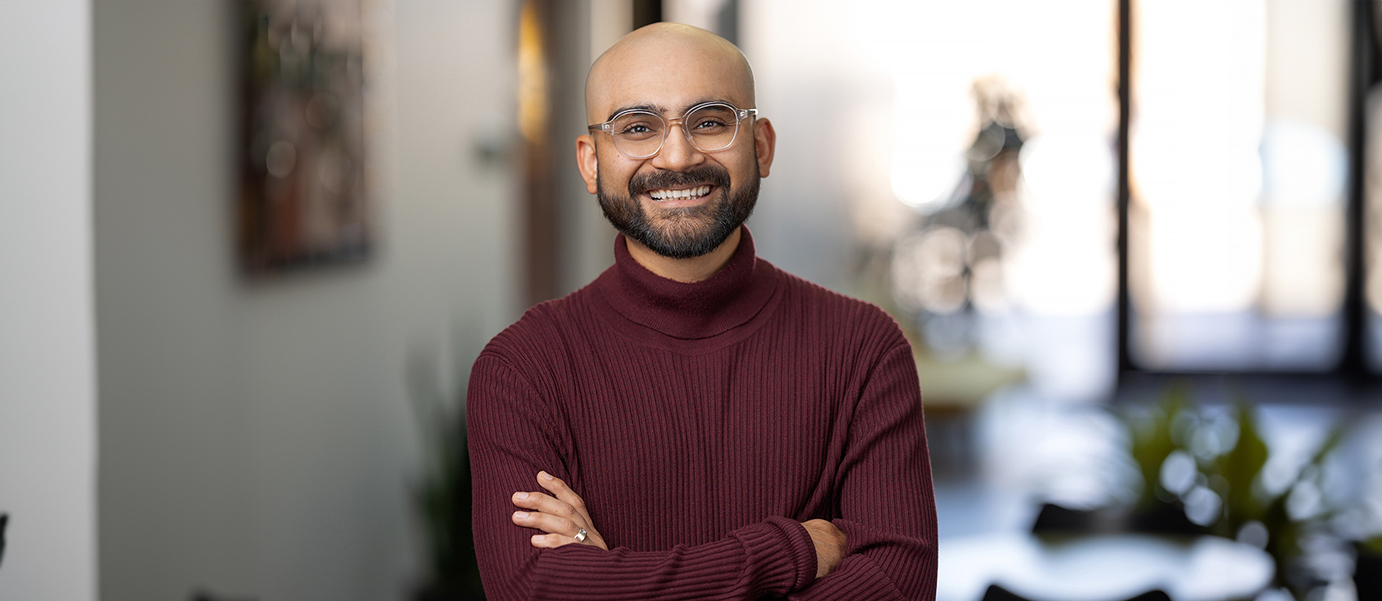 A smiling man with glasses and a beard stands indoors, arms confidently crossed in a maroon sweater, like he's the main navigation in his own story. The background is softly blurred.