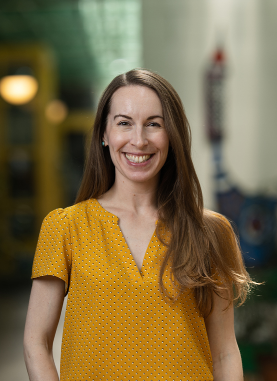 A woman with long hair smiles while wearing a yellow polka dot blouse. She's standing in an indoor setting, appearing nimble and lively, with blurred colorful objects in the background.