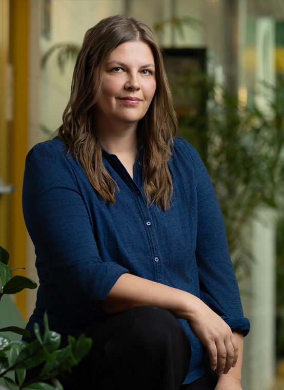 The nimble woman with long brown hair in a blue shirt sits indoors near plants, looking confidently at the camera, as if ready to lead her team.