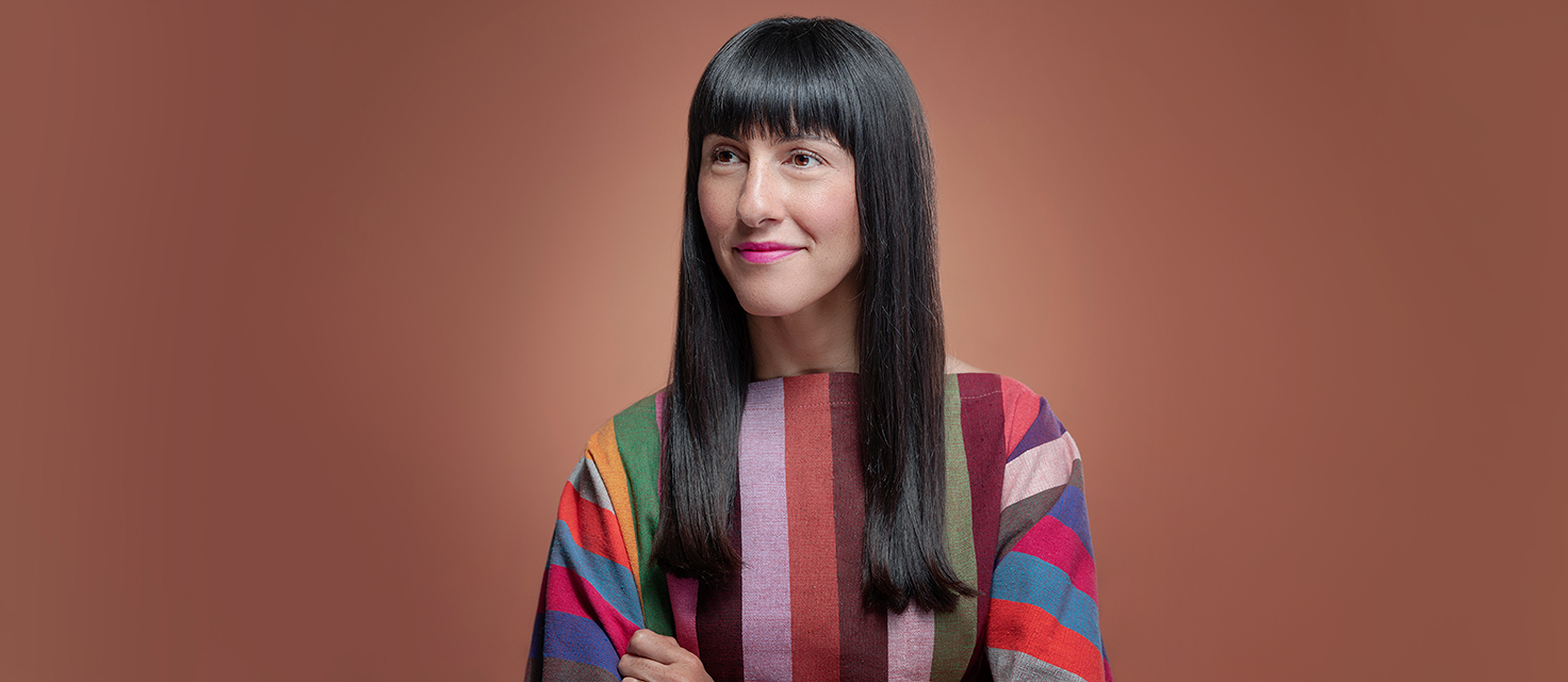 A person with long dark hair wearing a colorful, striped top is captured against a brown background, effortlessly drawing the eye like the main navigation of an intriguing webpage.