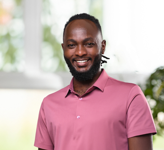 A person wearing a pink shirt smiles in a brightly lit room, appearing nimble among the lush green plants in the background.