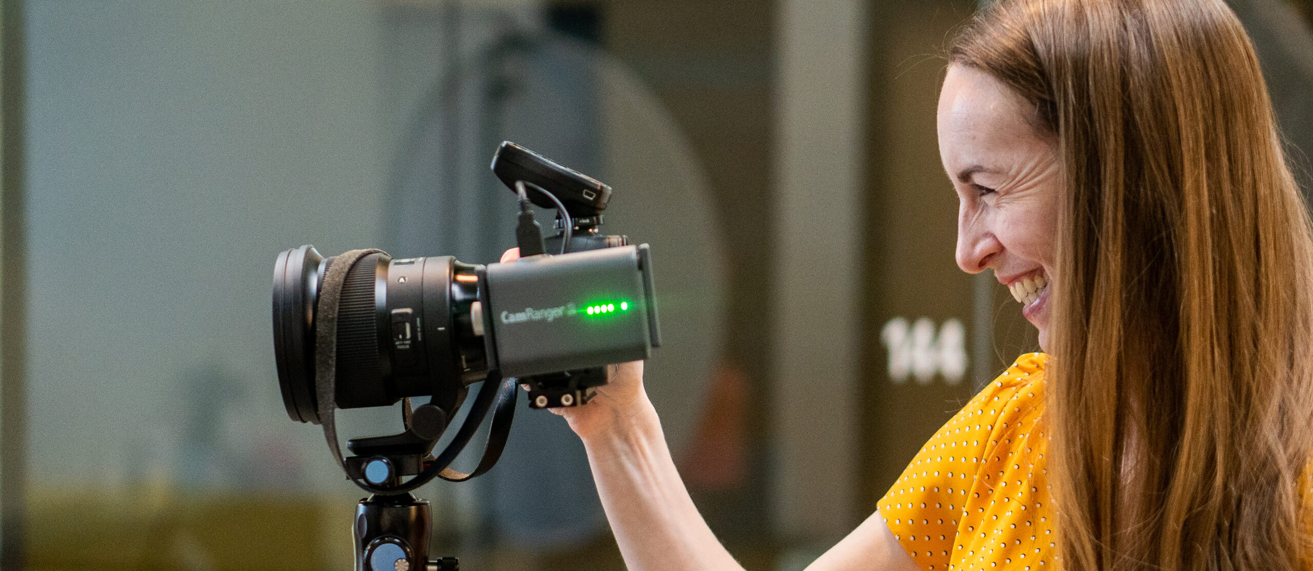 Woman with long hair smiles while adjusting a camera on a tripod indoors. She wears a yellow polka dot shirt, her eyes darting occasionally toward the main navigation board on the wall, ensuring everything is perfectly set for her shoot.