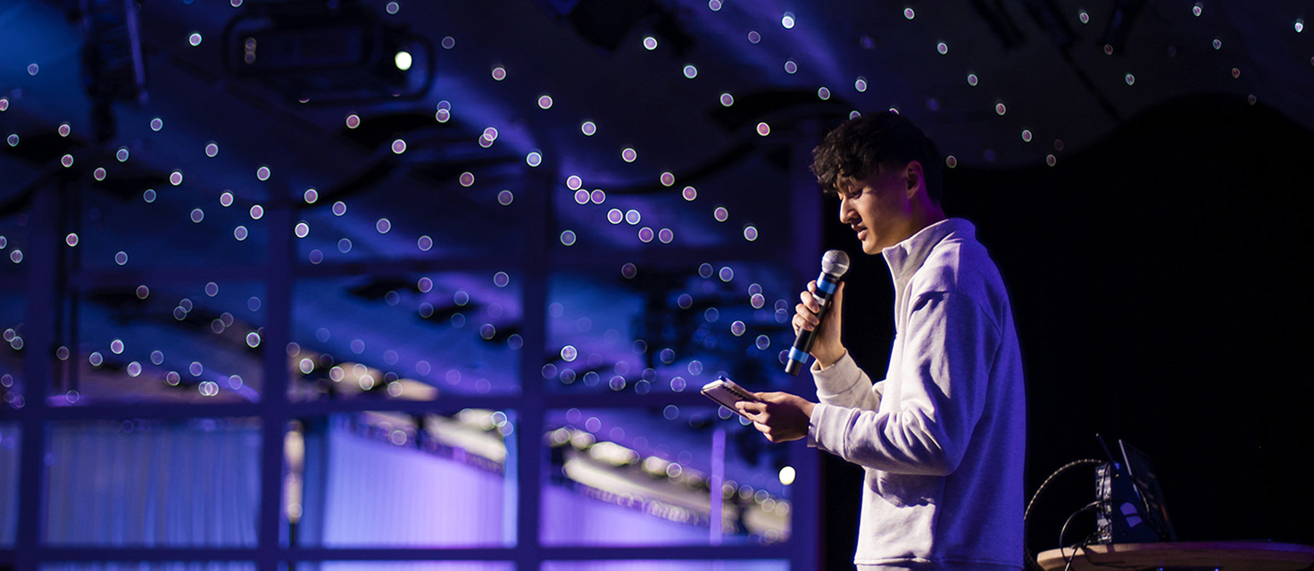 A person stands center stage, holding a microphone and phone, navigating their speech beneath a ceiling adorned with numerous small lights.