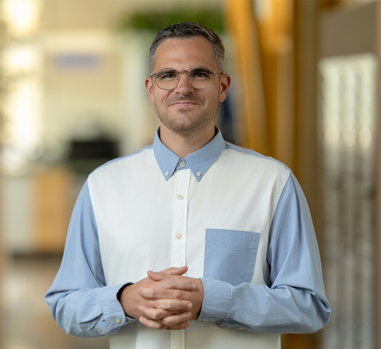 A person with short hair and glasses, wearing a blue and white collared shirt, stands indoors with their hands clasped, exuding a nimble confidence that's essential for any team.