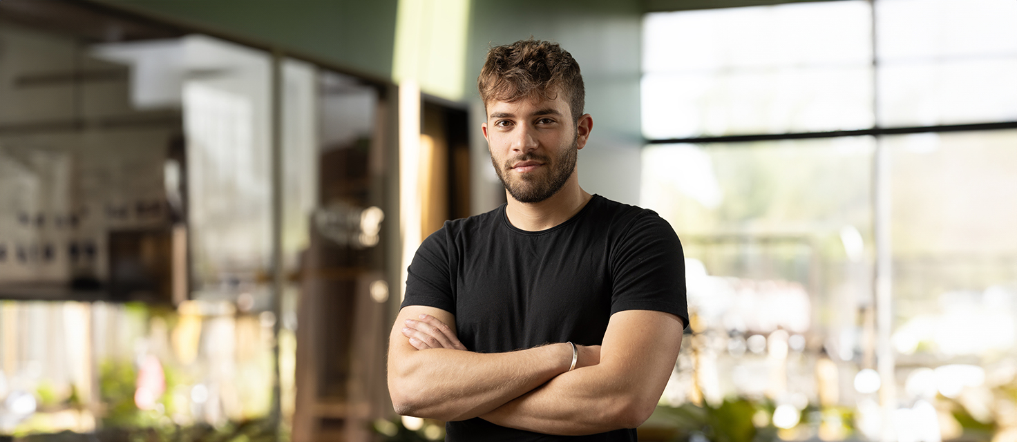 A person with short hair and a beard stands indoors, arms confidently crossed in a black t-shirt. Main Navigation is seamless through the blurred background of large windows lit by soft natural light.