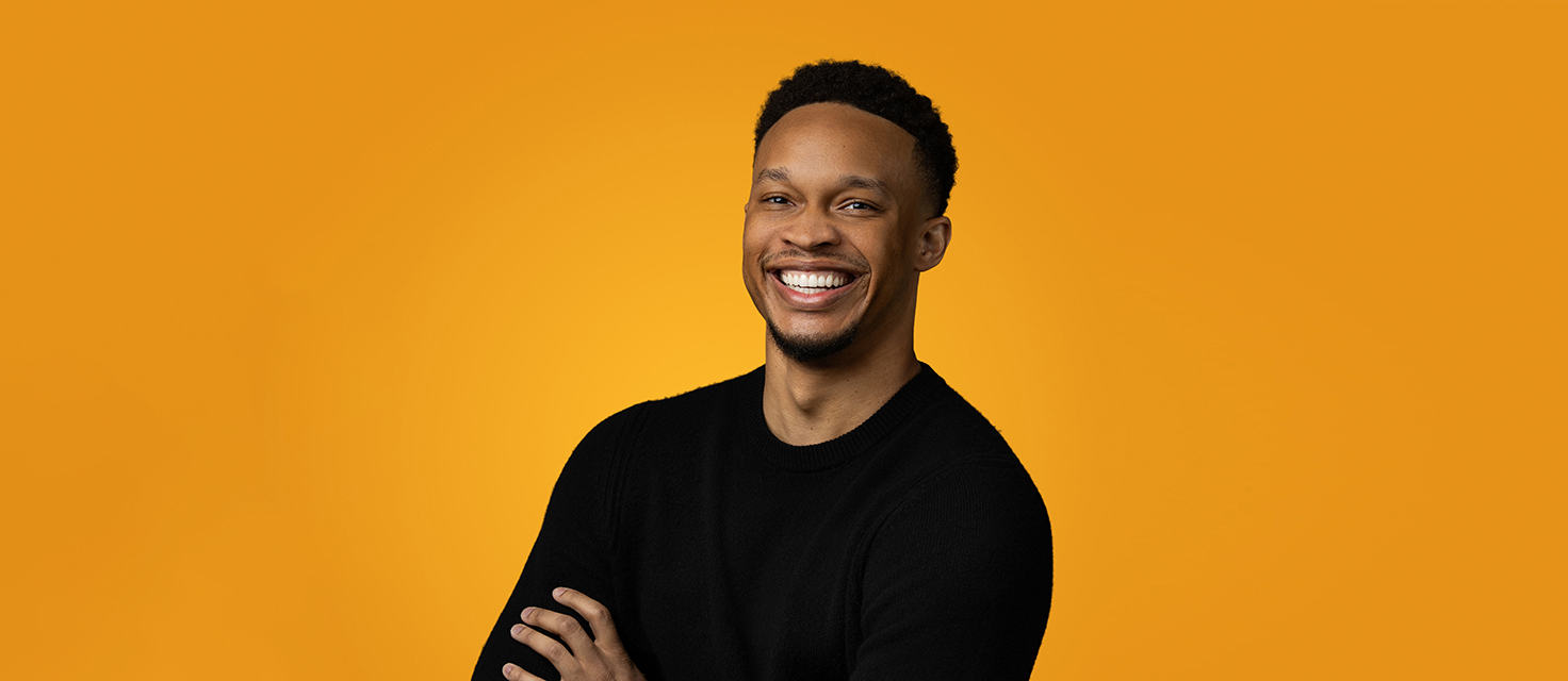 A smiling man in a black sweater stands with arms crossed against an orange background, exuding confidence and charisma, much like the main navigation of a well-designed website.