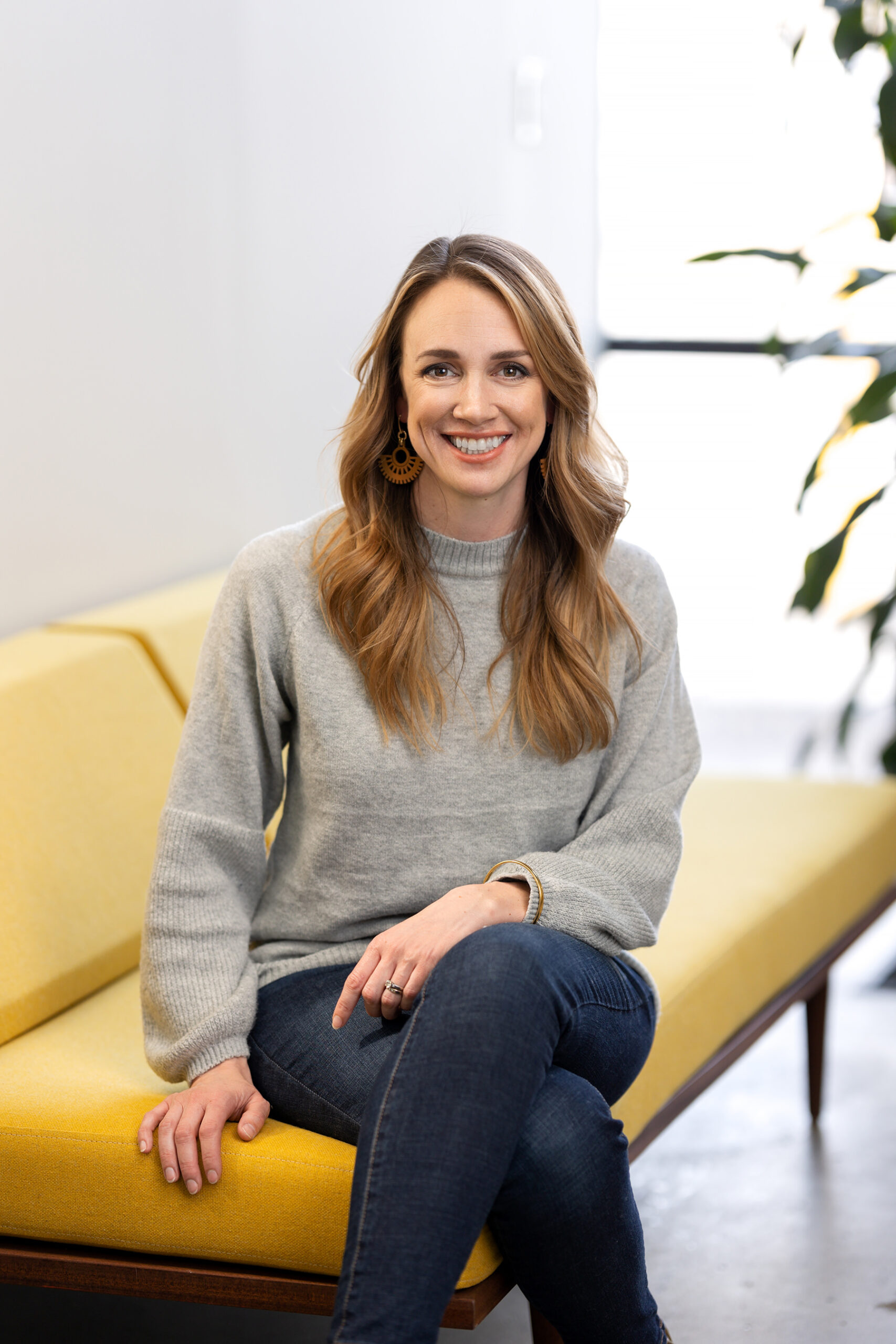 A smiling woman wearing a gray sweater and jeans sitting on a yellow couch for Denver headshots.