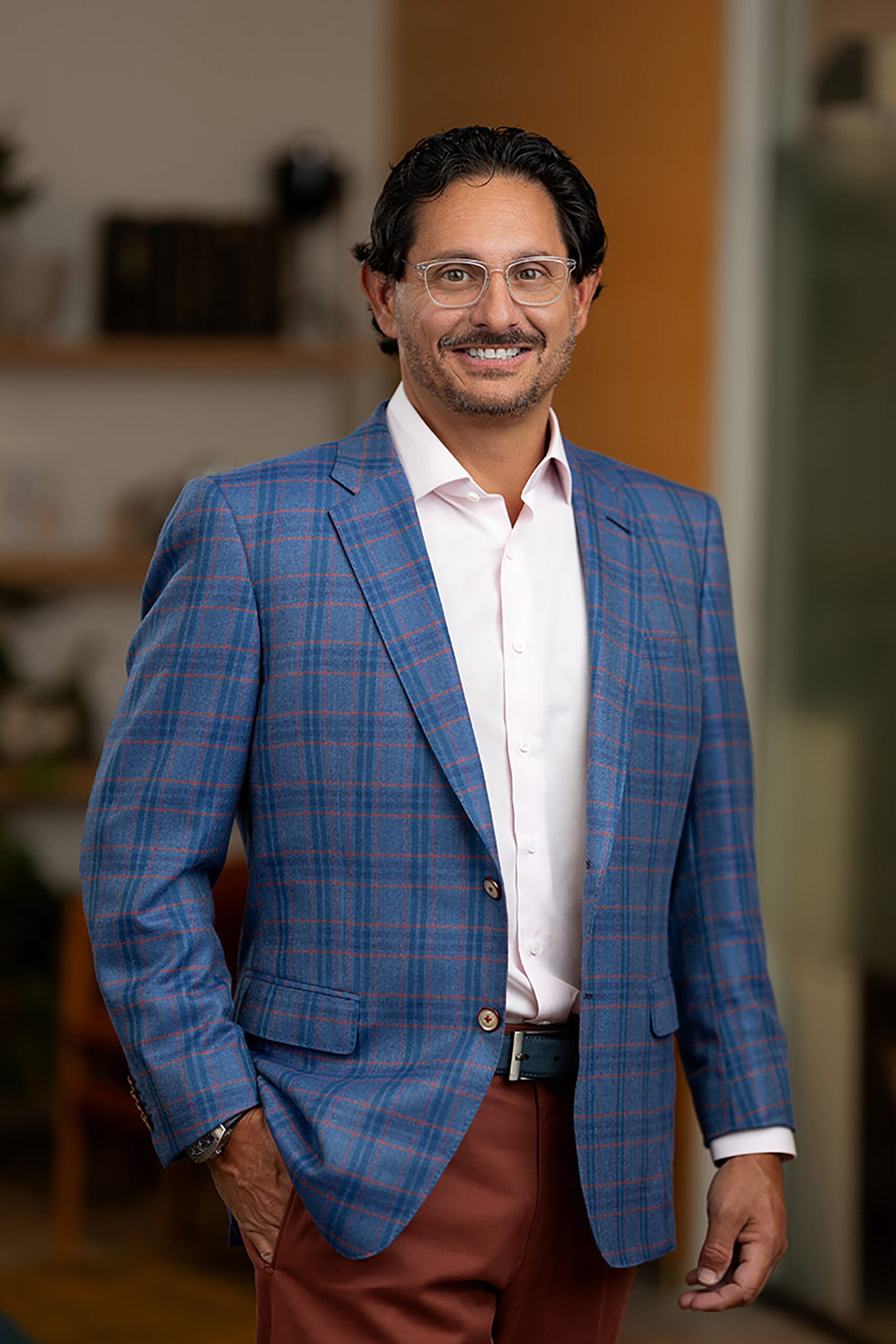 A man in a plaid blazer and brown trousers standing with one hand in his pocket, smiling at the camera for his Denver headshots.