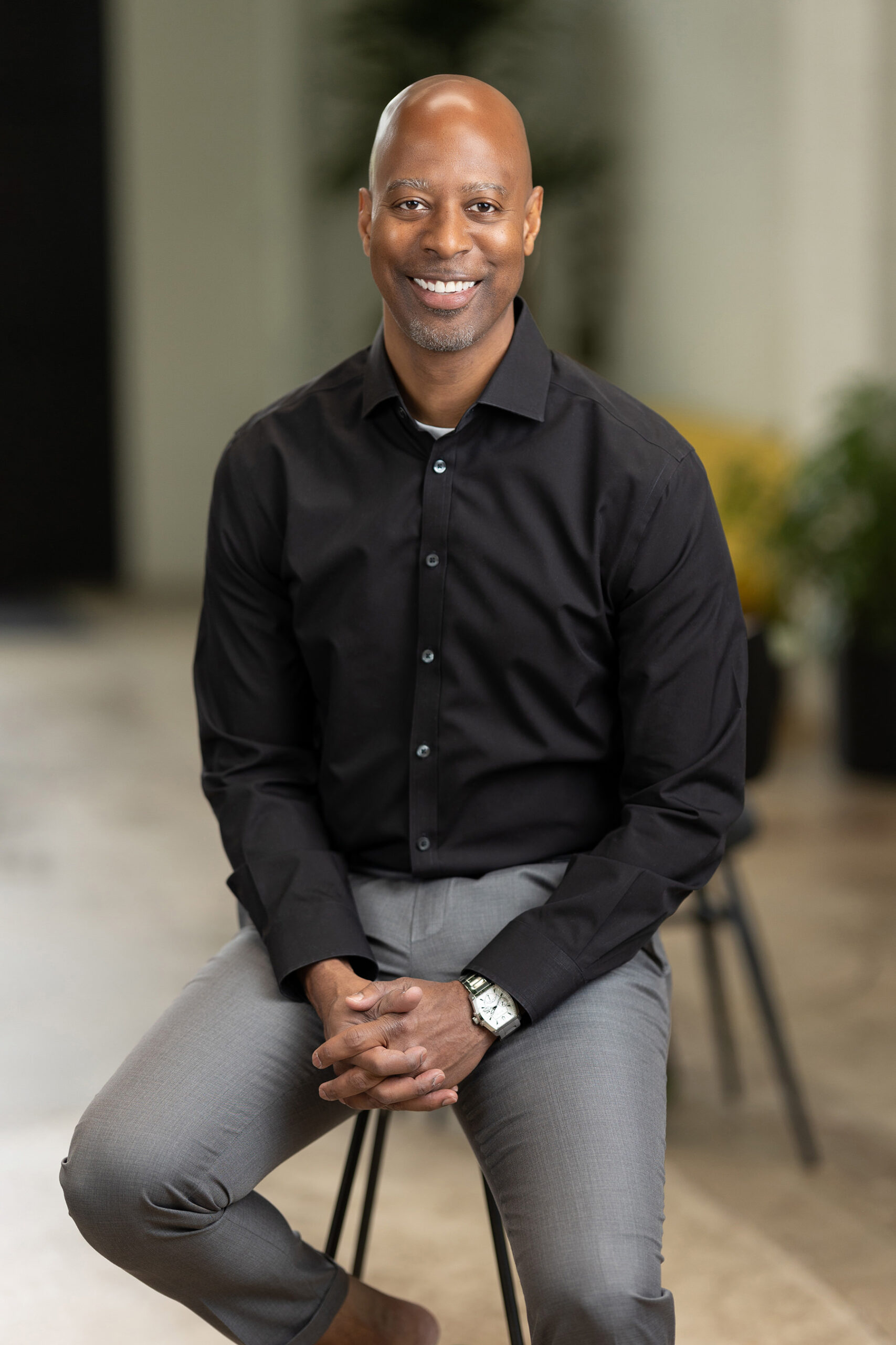 Man seated on a stool with a confident smile for Denver headshots, dressed in a black shirt and gray pants.