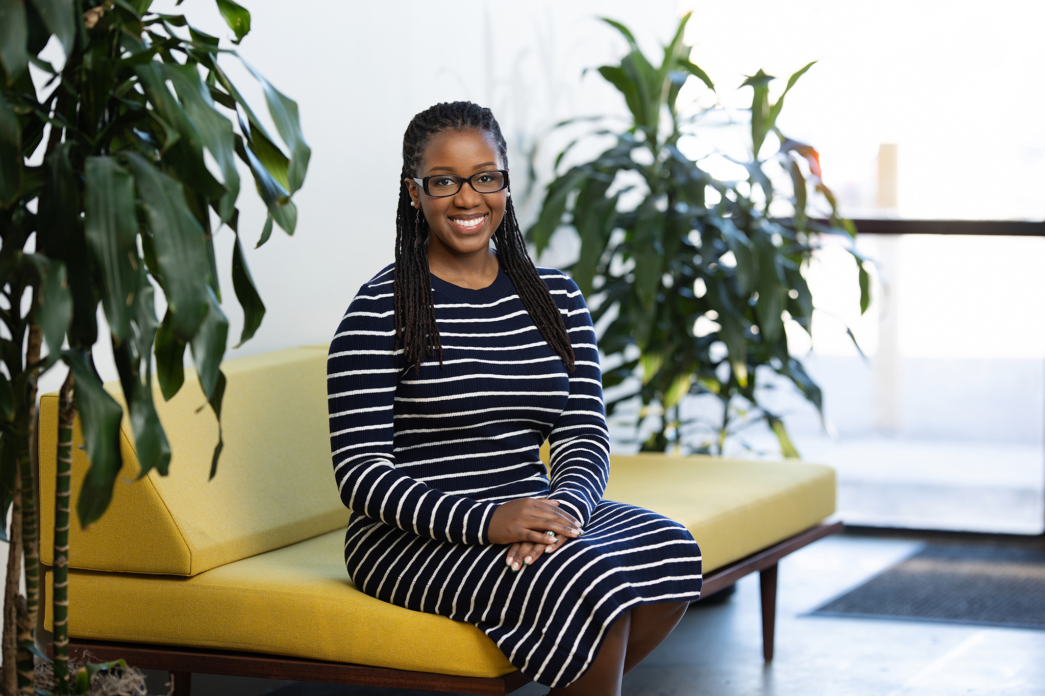 A woman in a striped dress smiling while sitting on a yellow couch, with plants in the background, poses for Denver headshots.