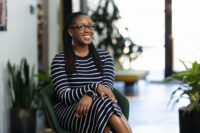 A smiling woman with glasses, wearing a striped dress, sitting in a green chair in a modern office space for her Denver headshots.