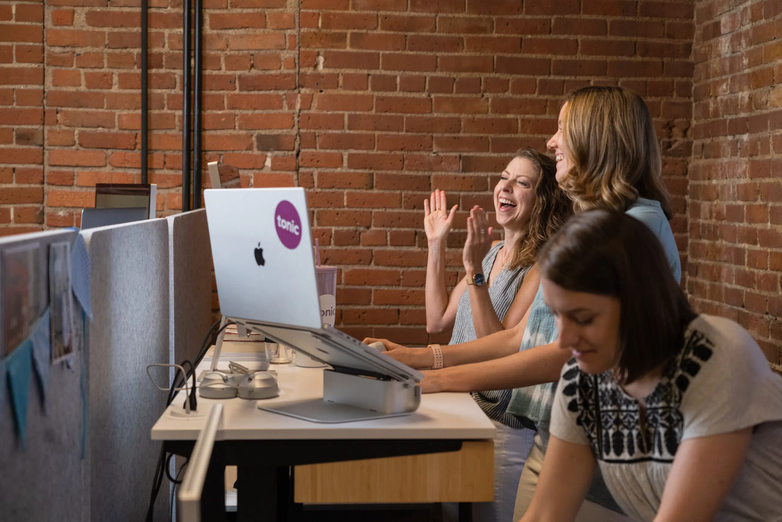 Three women working at standing desks in an office with brick walls. One woman in the middle is laughing and clapping while another types on a laptop. The third woman, with a focused expression, epitomizes the professionalism you'd expect from Denver Headshot Co.