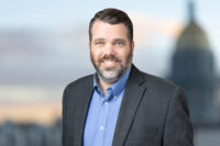 Man with dark hair and beard wearing a blue shirt and gray blazer, standing in front of a blurred background featuring a dome structure; a professional capture by Denver Headshot Co.