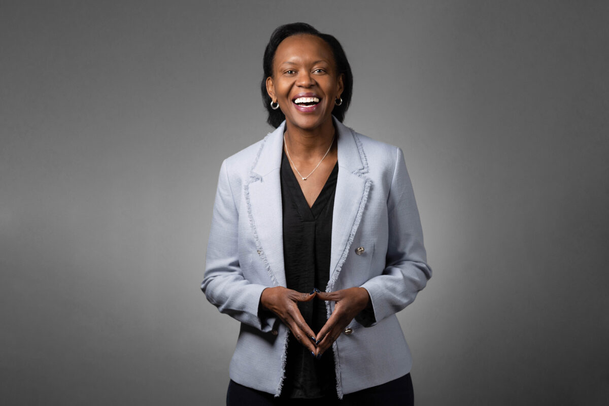 A woman in a light blue blazer and black top is smiling and standing against a plain gray background with her hands loosely clasped, showcasing the professional quality of Denver Headshot Co.