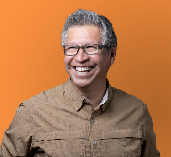 A person with gray hair and glasses, wearing a beige shirt, smiles against an orange background in this stunning Denver Headshot Co capture.