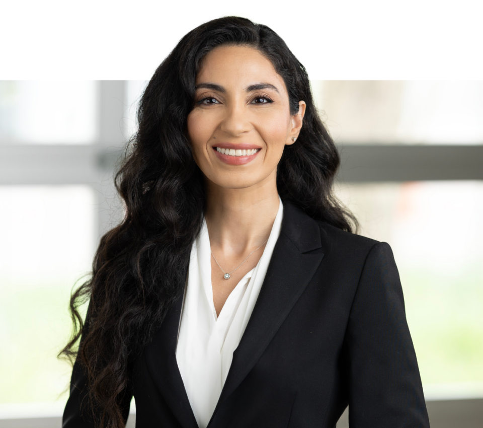 A woman with long, dark hair is standing, smiling, and wearing a black blazer over a white blouse. The background appears to be indoors and is slightly blurred. This professional portrait was expertly captured by Denver Headshot Co.