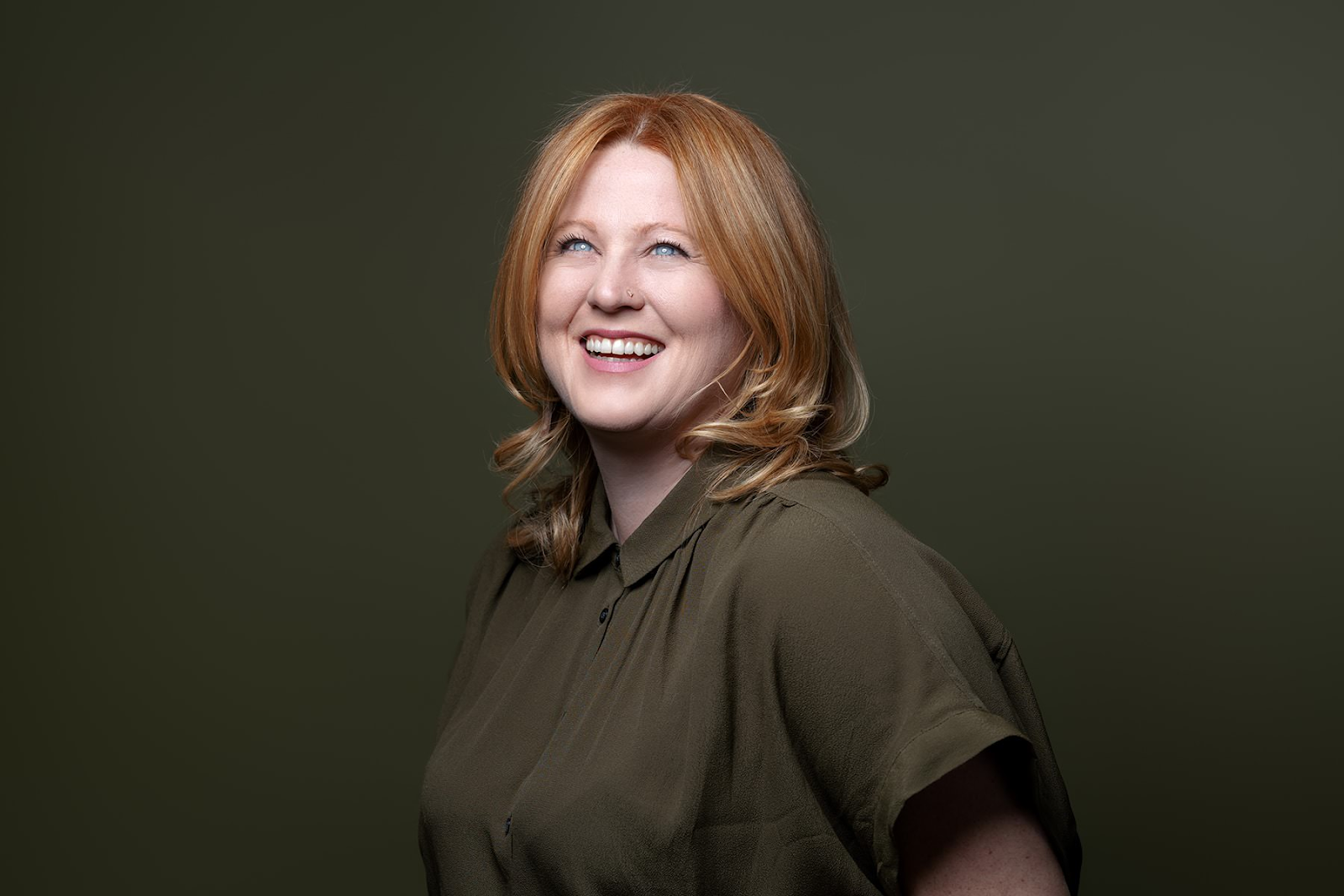 A woman with red hair and wearing a green shirt smiles while looking upward, the perfect capture by Denver Headshot Co, set against a dark green background.