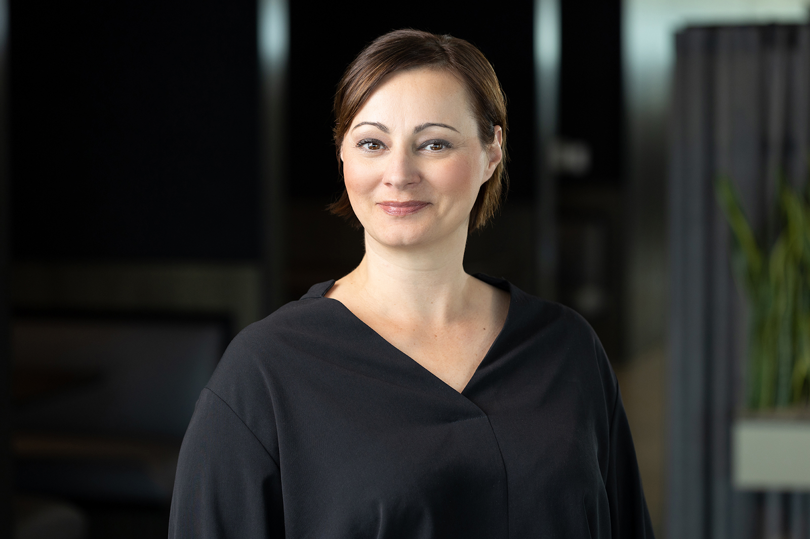 A person with short brown hair, dressed in a black top, stands indoors with a neutral expression and plants in the background—a perfect subject for Denver Headshot Co.