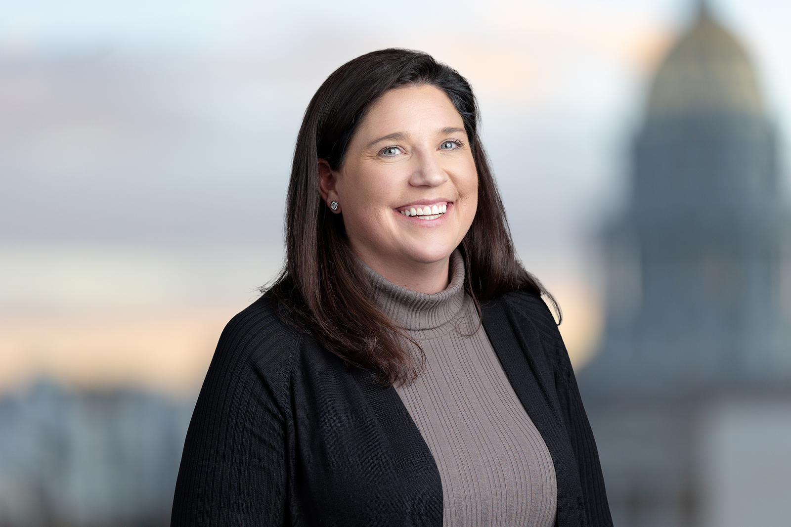 A woman with long brown hair, wearing a grey turtleneck and black sweater, smiles against a blurred background featuring a domed building, captured perfectly by Denver Headshot Co.