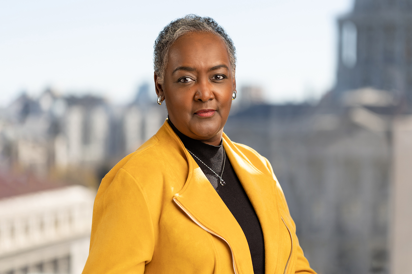 A woman with short gray hair is wearing a yellow jacket and a black top while standing outdoors, with Denver's cityscape in the blurred background—captured perfectly by Denver Headshot Co.