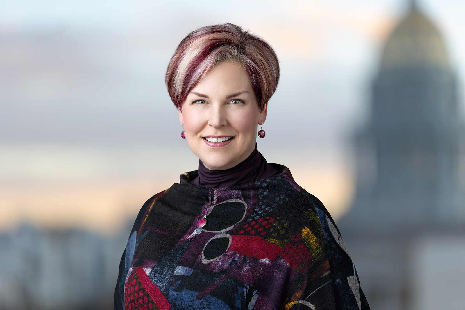 A woman with short, styled hair, wearing a colorful shawl, stands smiling in front of a blurred background featuring a domed building—perfect for a Denver Headshot Co portfolio.