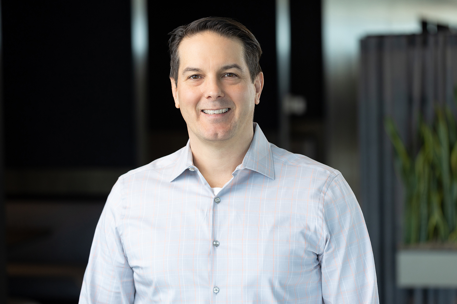 A man in a light-colored button-up shirt stands indoors with a neutral expression, captured flawlessly by Denver Headshot Co. The background is blurred with dark and light areas.