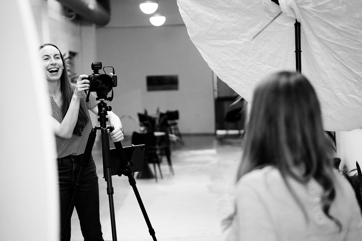 A woman from Denver Headshot Co is holding a camera and laughing while taking a photo of another person in a studio setting.