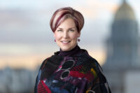 A woman with short, highlighted hair wearing a colorful patterned top smiles with a blurred domed building in the background, capturing the spirit of a Denver Headshot Co session.