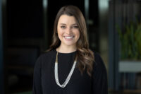 A woman with long hair, wearing a black top and a layered necklace, is smiling against a blurred indoor background. Captured by Denver Headshot Co, her confident expression shines through the lens.