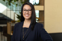 A woman with long dark hair and glasses is smiling and wearing a dark blazer and necklace, standing in an office environment. Captured by Denver Headshot Co, her professional demeanor shines through.