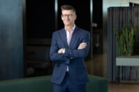 A man in a blue suit with glasses stands confidently with his arms crossed in an office setting, captured perfectly by Denver Headshot Co.