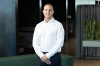 A man in a white shirt stands indoors, smiling at the camera with hands clasped in front. The background includes office furniture and a plant, perfectly captured by Denver Headshot Co.