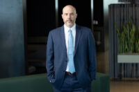 A man in a blue suit and light blue tie stands indoors, perfectly posed for Denver Headshot Co. Behind him, the background features dark walls, a green plant, and subtle lighting.