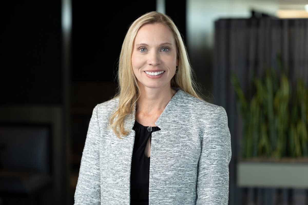 A woman with long blonde hair, wearing a light gray blazer over a black top, is smiling warmly while standing indoors in front of a blurred background; this Denver Headshot Co portrait truly captures her personality.