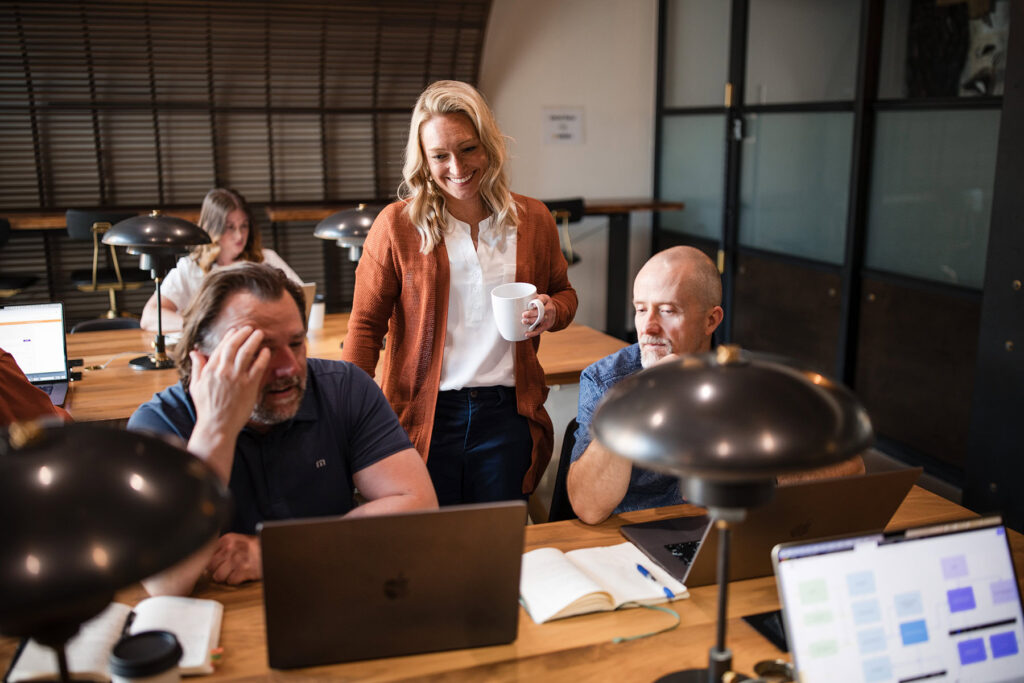 A woman stands holding a cup and smiles while engaging with two men seated at a table with laptops in a communal workspace, perfectly capturing the collaborative spirit that Denver Headshot Co is known for.