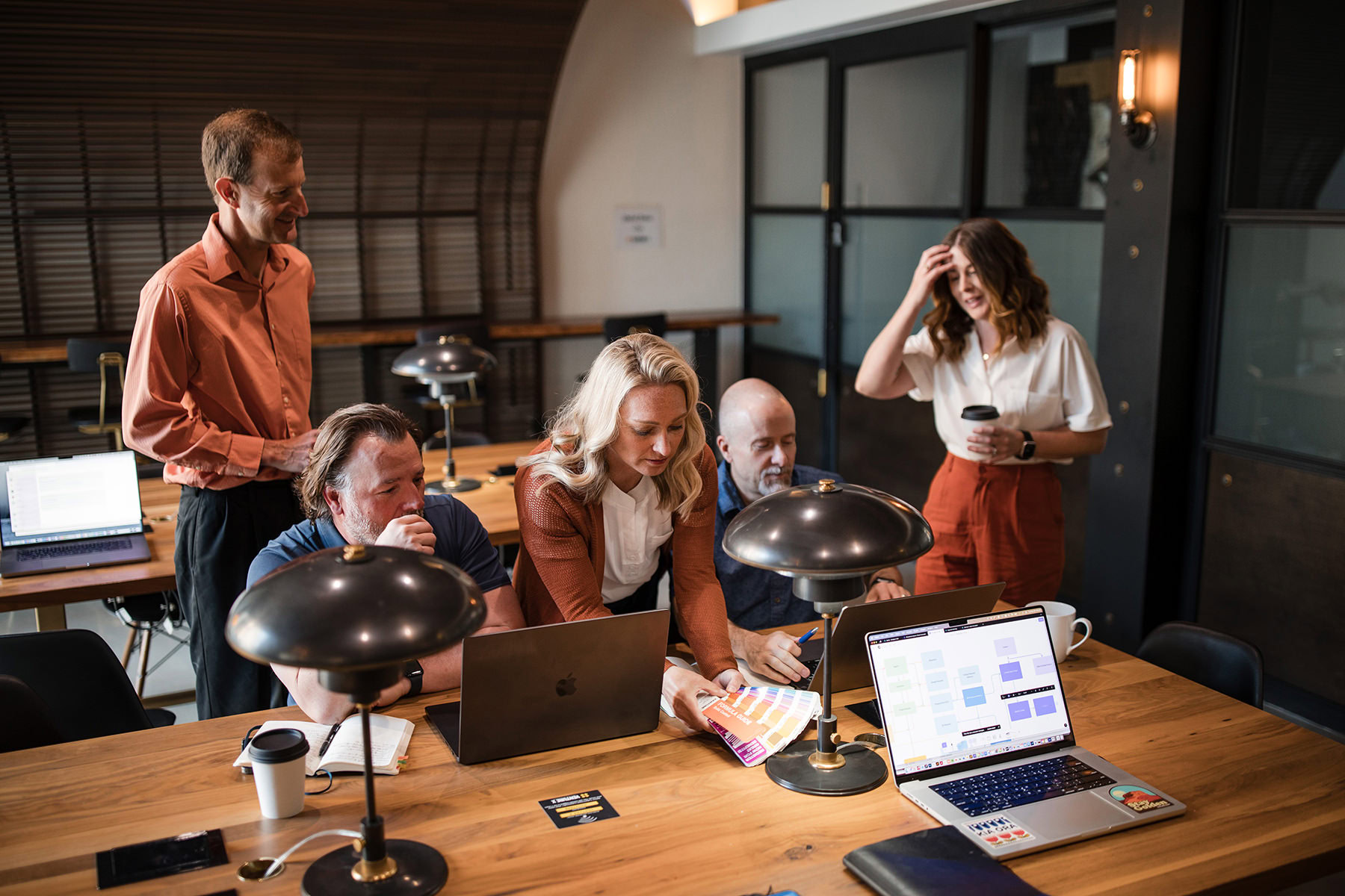 A group of five people in a meeting room are gathered around a table with laptops and documents, engaging in a discussion. One person is standing with a Denver Headshot Co mug, three are seated, and another is presenting ideas on their laptop.