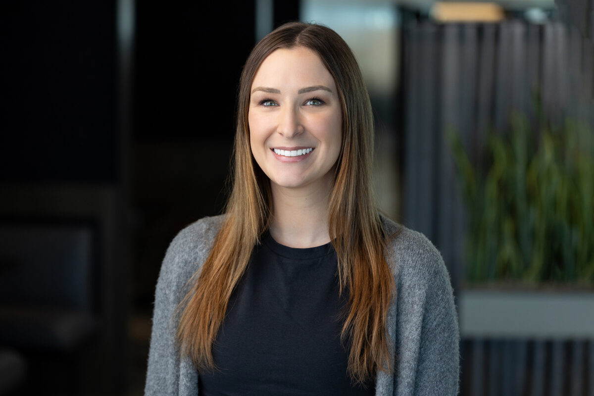 A person with long brown hair and a gray sweater smiles while standing indoors, perfectly captured by Denver Headshot Co.