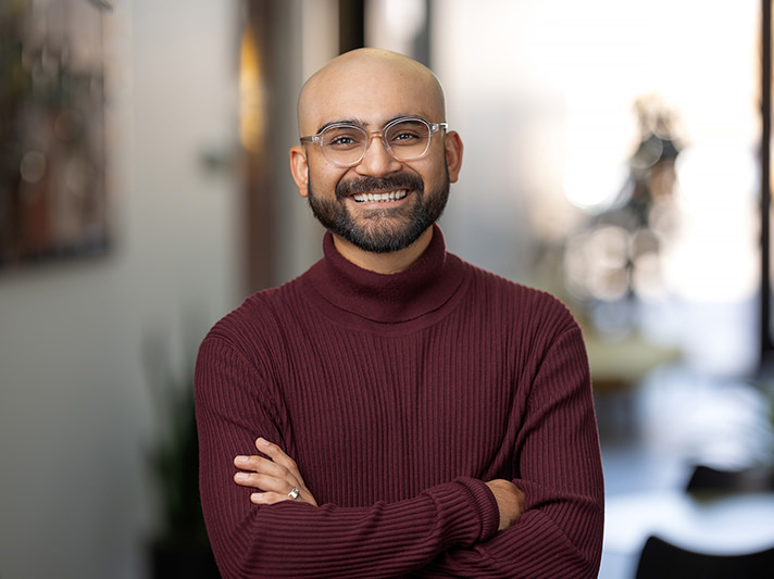 A person with glasses and a beard stands indoors, smiling with arms crossed, wearing a maroon sweater, perfectly captured by Denver Headshot Co.