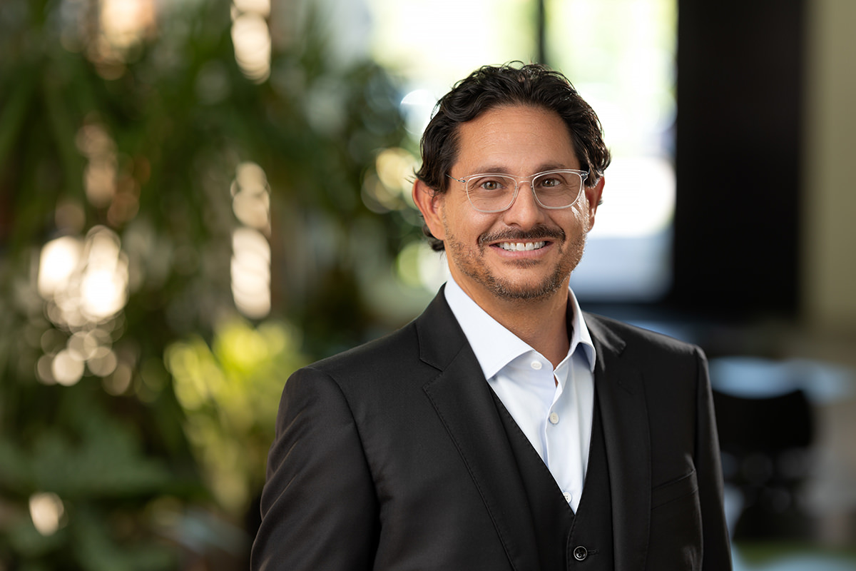 A man with glasses and short, dark hair wearing a suit smiles in an indoor setting, captured beautifully by Denver Headshot Co, with blurred greenery in the background.