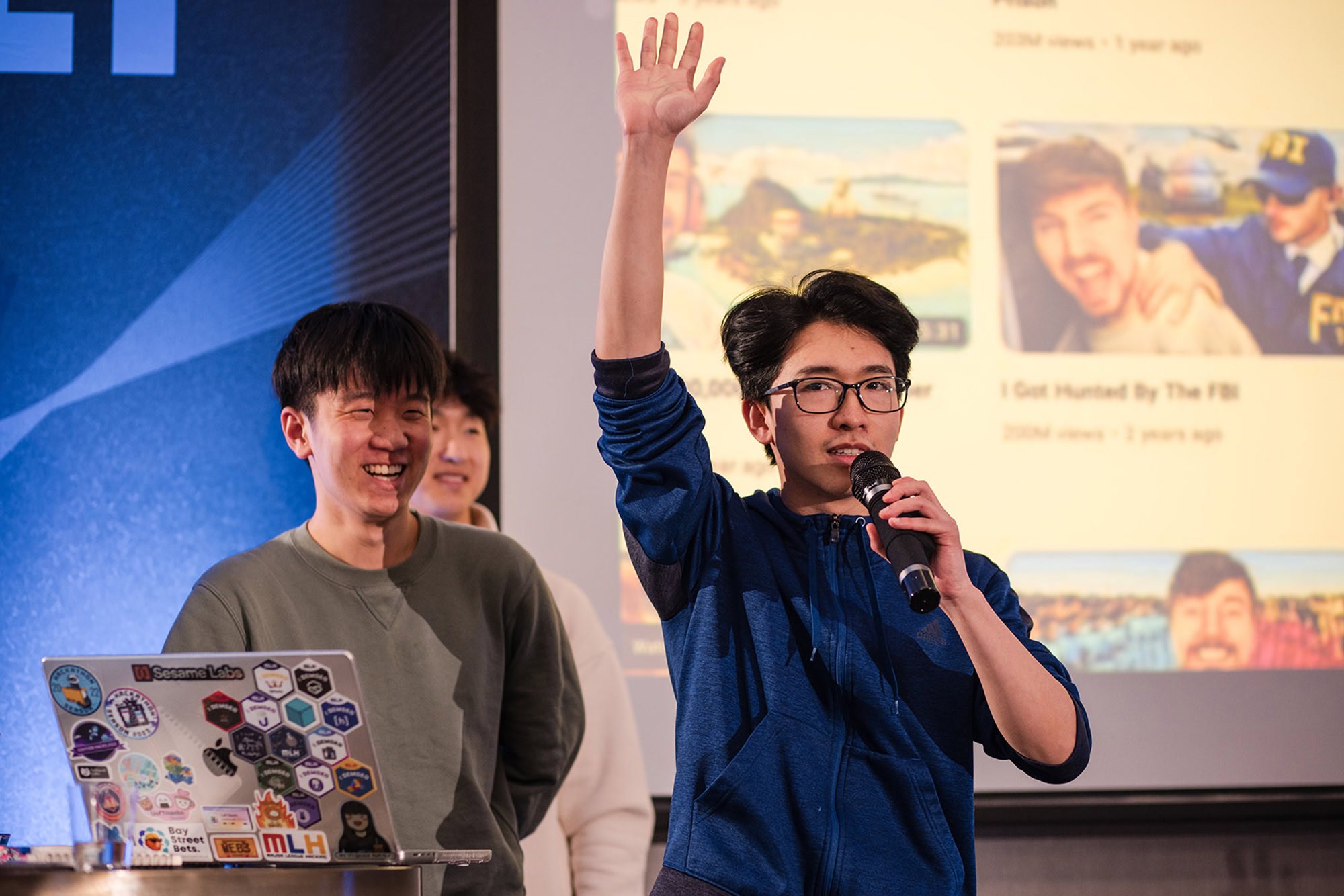 Two people are on stage presenting. One is speaking into a microphone and raising a hand, while the other is smiling and standing near a laptop covered in Denver Headshot Co stickers. A large screen displays videos in the background.