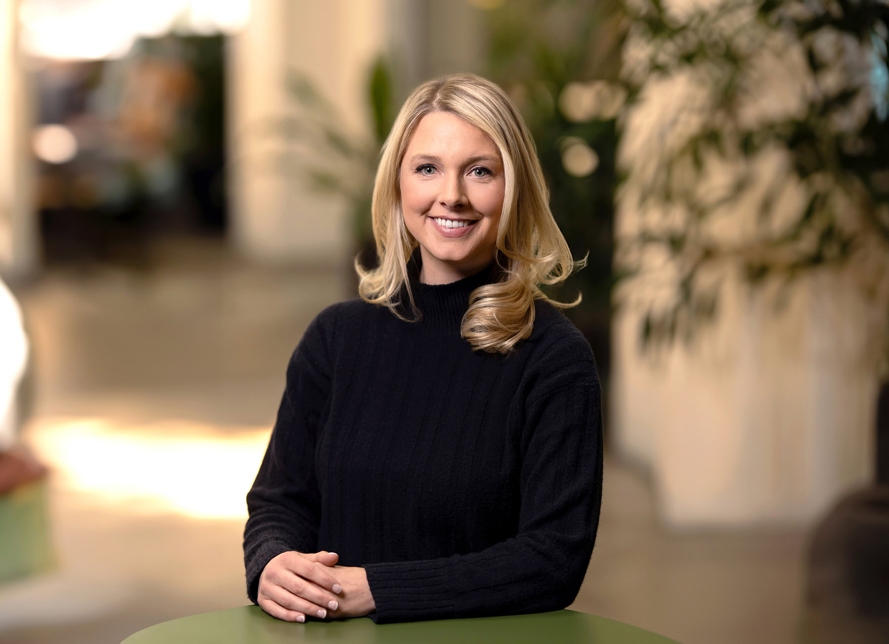 A woman with shoulder-length blonde hair, wearing a black sweater, smiles at the camera while sitting at a table. The indoor background features blurred plants and a hallway, capturing the warmth and professionalism typical of Denver Headshot Co.