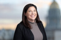 A woman with long brown hair, smiling, wearing a gray turtleneck and black jacket, stands against a blurred background of a domed building and sky at sunset. Captured by Denver Headshot Co.