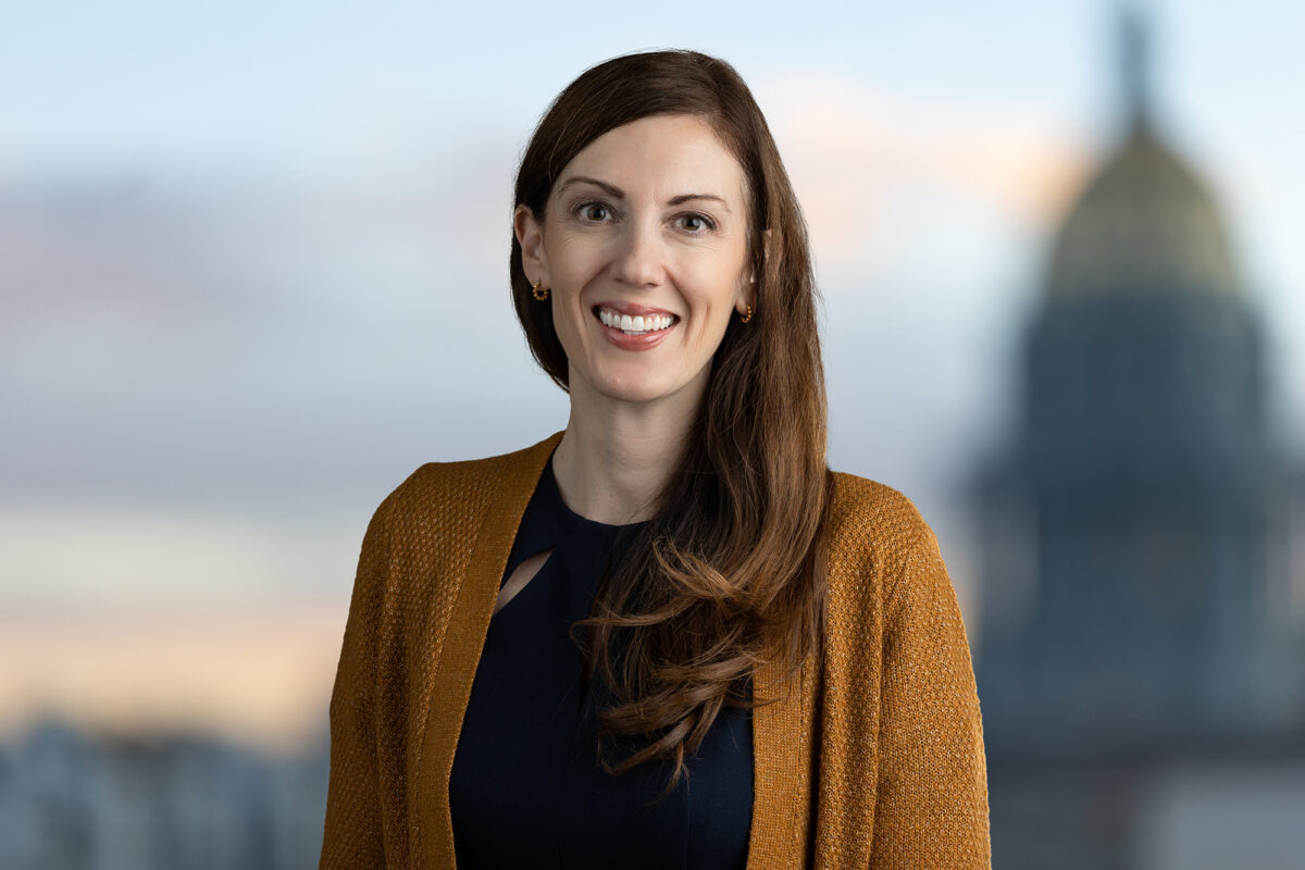 A woman with long brown hair, wearing a black top and mustard cardigan, is smiling at the camera with a blurred building in the background, courtesy of Denver Headshot Co.