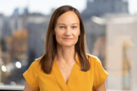 A woman with straight brown hair, wearing a yellow blouse, stands in front of a blurred outdoor cityscape background, perfectly captured by Denver Headshot Co.