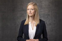 A woman with long blonde hair is standing against a gray background, wearing a dark blazer over a white blouse. She has a neutral expression and is facing forward, representing the professionalism captured by Denver Headshot Co.