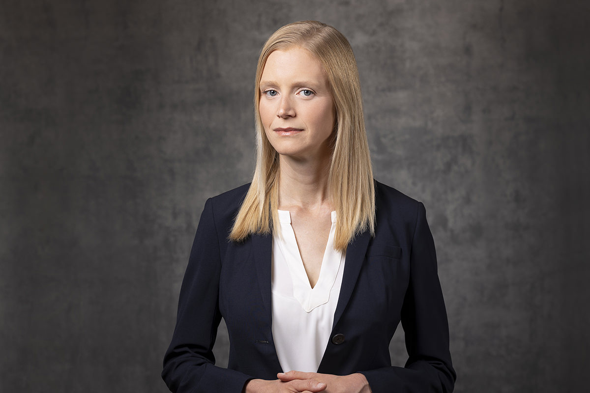 A woman with long blonde hair is standing against a gray background, wearing a dark blazer over a white blouse. She has a neutral expression and is facing forward, representing the professionalism captured by Denver Headshot Co.