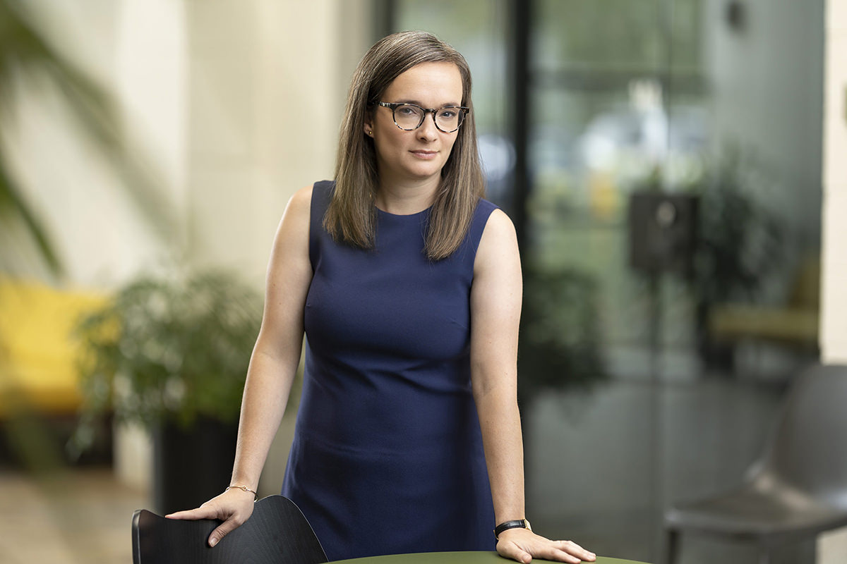 A woman with long hair and glasses, wearing a navy blue sleeveless dress, stands leaning on a table in a modern indoor setting with plants in the background, as captured by Denver Headshot Co.