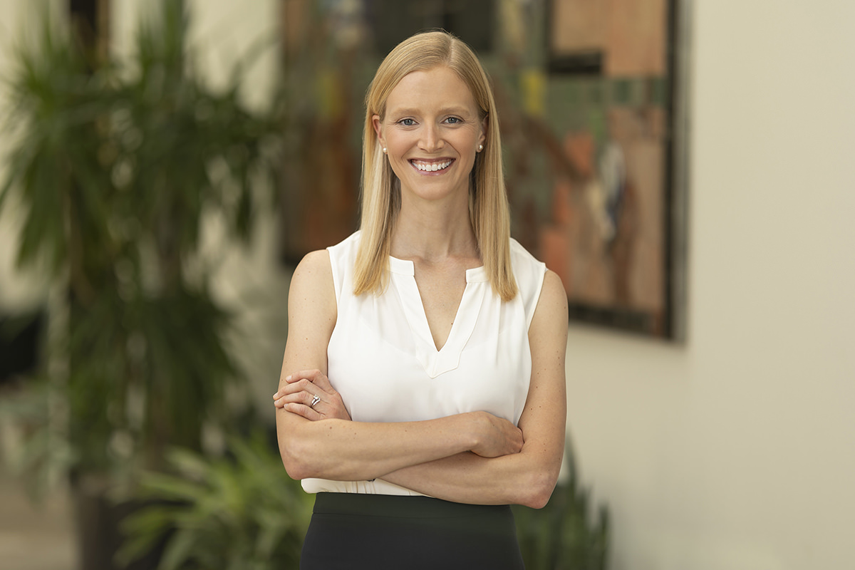 A woman with long blonde hair and a white sleeveless blouse stands indoors with her arms crossed, smiling at the camera. Behind her, plants and a colorful wall hanging add charm to the scene captured perfectly by Denver Headshot Co.