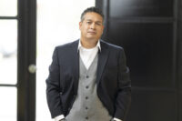 A man in a dark suit jacket and gray vest stands indoors, looking at the camera with his hands in his pockets, showcasing the impeccable work of Denver Headshot Co.